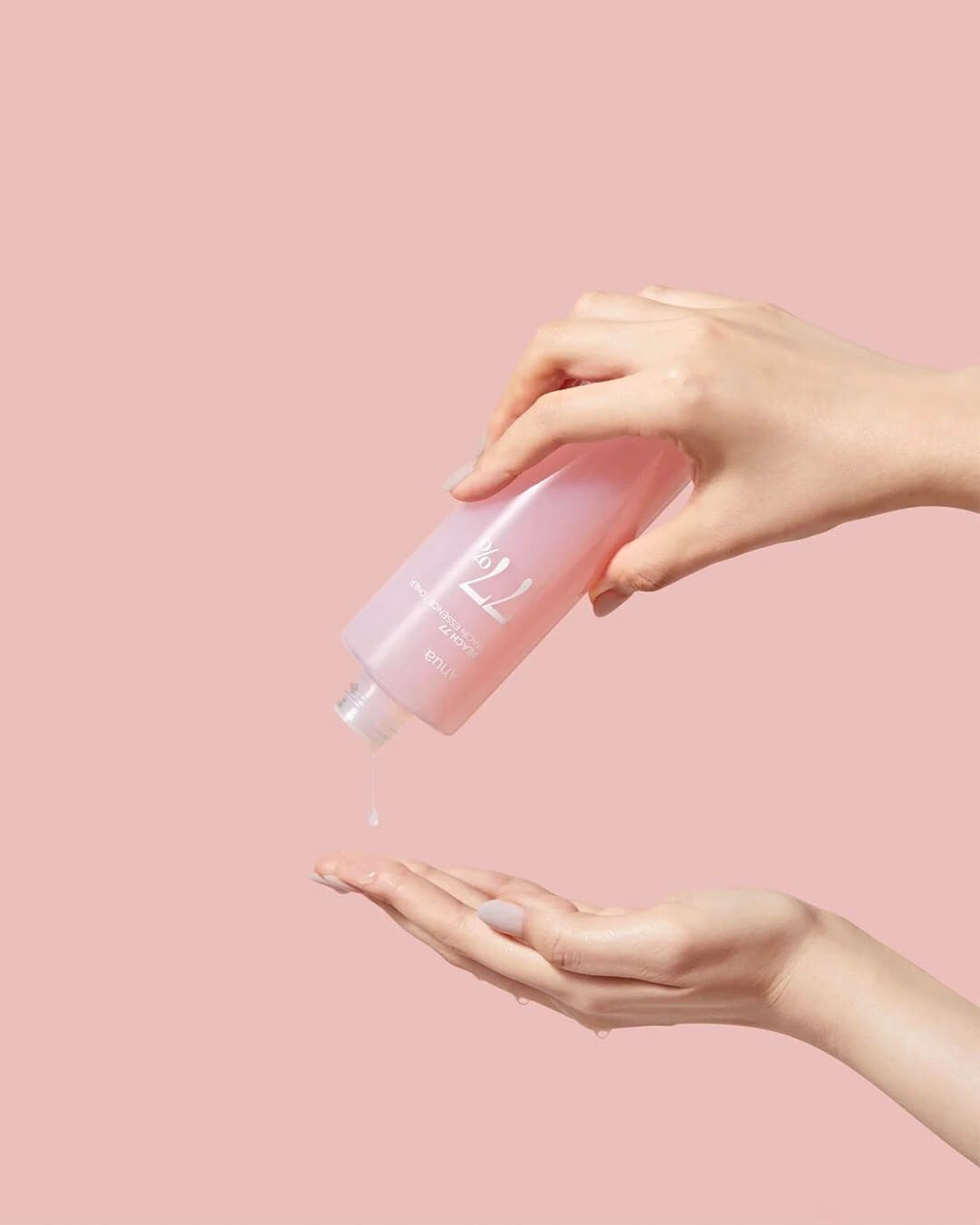 Hand holding a pink bottle with a pump, dispensing liquid onto another hand against a pink background